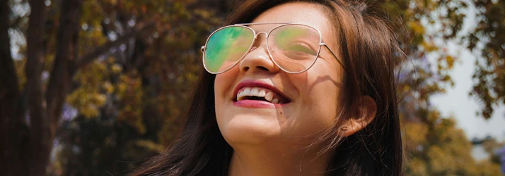 A smiling woman wearing reflective sunglasses, showing white teeth and pink lipstick, enjoying a sunny day outdoors.