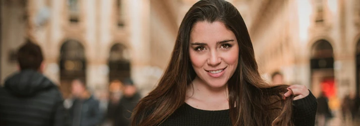 A woman with long brown hair smiles confidently at the camera, standing in a blurred urban setting.